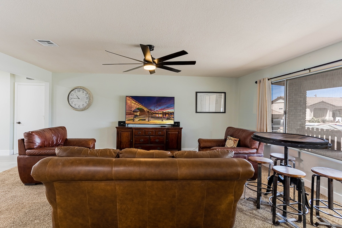 The living area showcases a comfortable leather sofa and armchairs, oriented towards a wall-mounted smart TV. A ceiling fan is present overhead, and large windows provide natural light, while a round bar table with stools adds to the casual dining space.