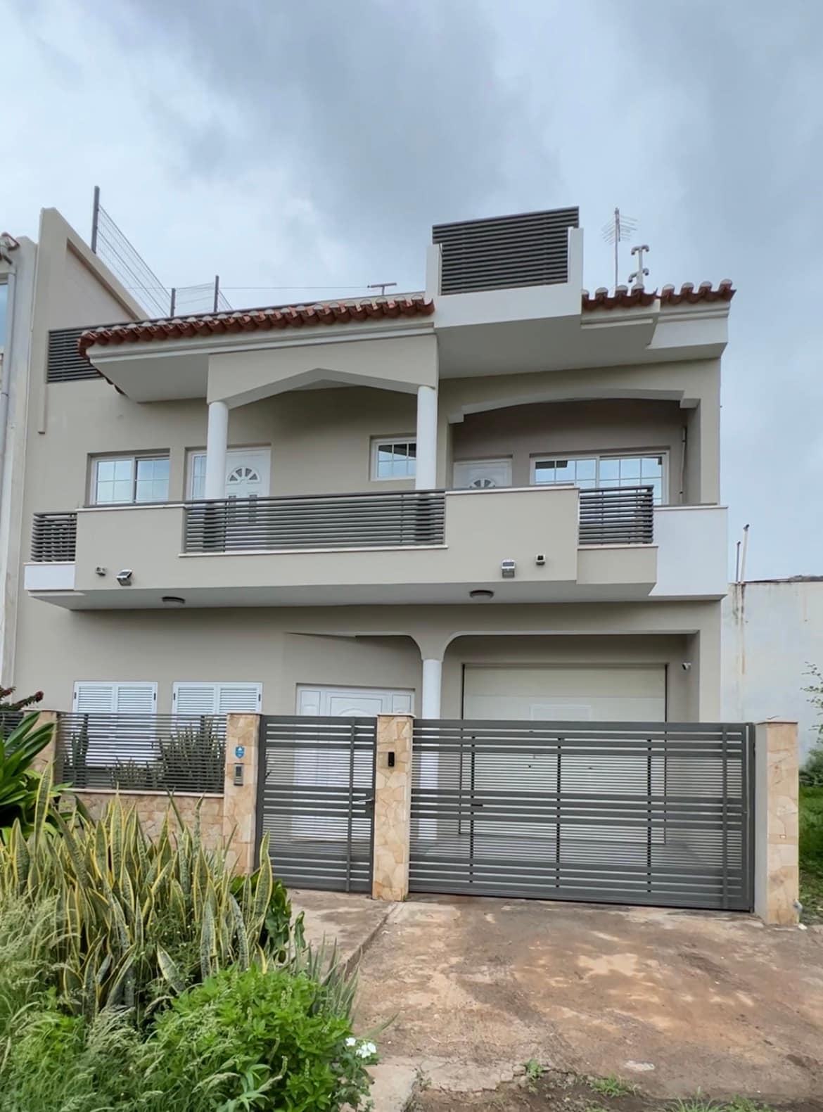 The exterior of a modern duplex house is displayed, featuring a light grey facade and a decorative balcony with a tiled roof. A secure gate marks the entrance, and landscaped greenery enhances the outdoor space, contributing to a welcoming environment.