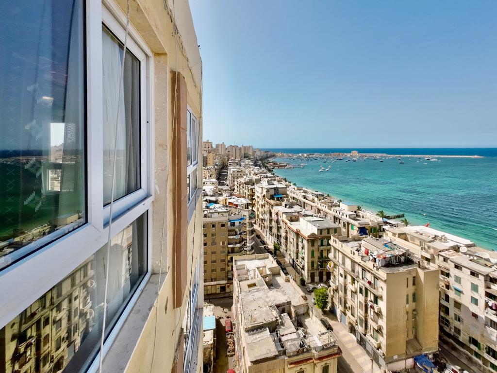 A panoramic view features the vibrant cityscape of Alexandria, with buildings lining the coast. The reflective glass window captures the clear blue sky and sparkling sea, creating a sense of openness and connection to the surrounding environment.