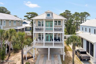 The home is showcased from the front, displaying a three-story structure with multiple balconies. Lush palm trees frame the entrance, and a paved area is visible beneath the house. The exterior features light siding and a metal roof, highlighting the coastal architecture.