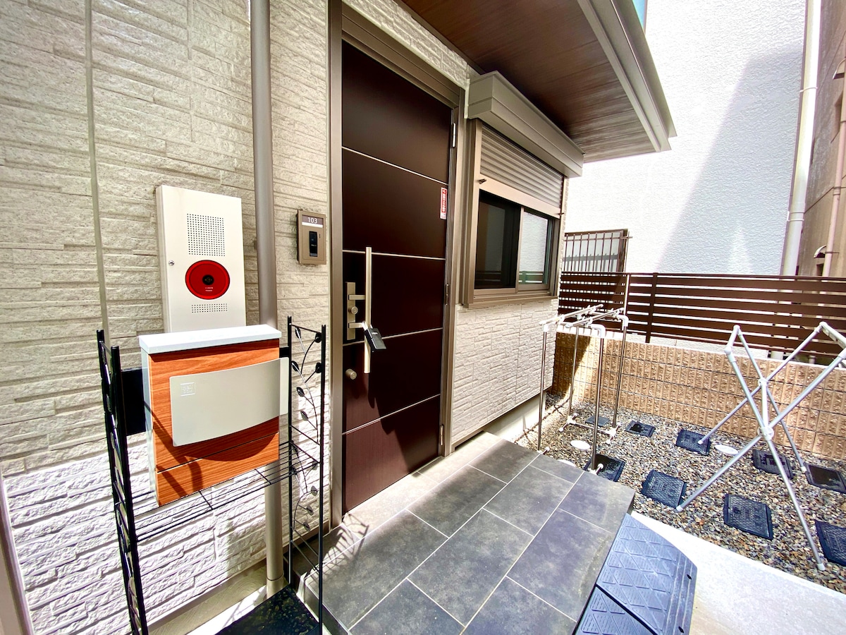 The exterior entrance features a modern door with a keypad and intercom system. A small mail slot is positioned above a wire rack. The area is surrounded by textured stone walls and includes a gravel space with clothes drying racks on the right.
