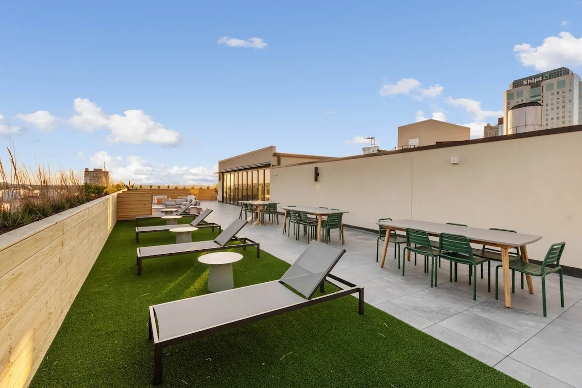 A rooftop lounge area is highlighted, featuring several sun loungers and tables surrounded by greenery. The space includes dining tables and modern chairs, offering views of the city skyline against a clear blue sky.