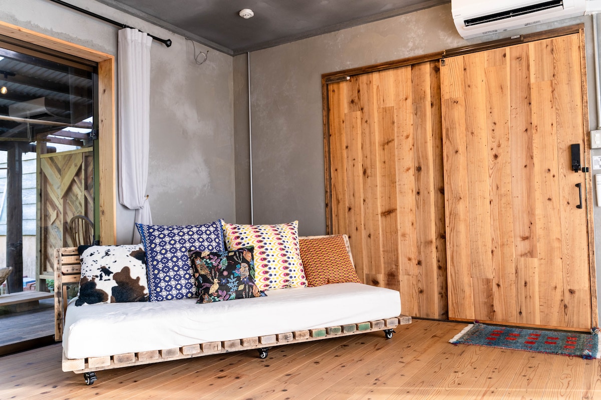 A spacious seating area features a low-profile sofa made from reclaimed materials, adorned with a variety of colorful patterned cushions. Large wooden sliding doors lead to an outdoor space, with natural light illuminating the warm wooden floor.