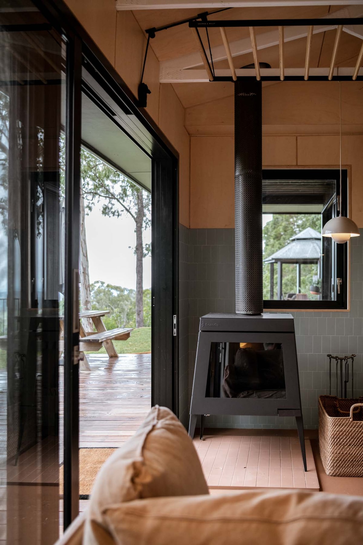 A modern interior space showcases a sleek black fireplace as the focal point. Large glass doors open to a wooden deck area, blending indoor comfort with outdoor scenery. Natural light fills the room, highlighting the clean lines and minimalist design.