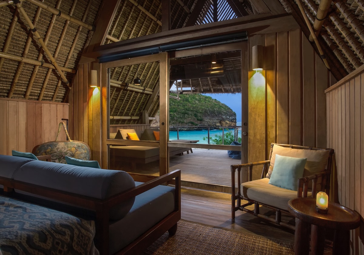 Inside view of a rustic beach hut featuring wooden walls and thatched ceilings. A spacious bed is adorned with soft blue linens, while a coffee table and chairs offer a cozy seating area. Open doors lead to a large terrace with stunning ocean views.