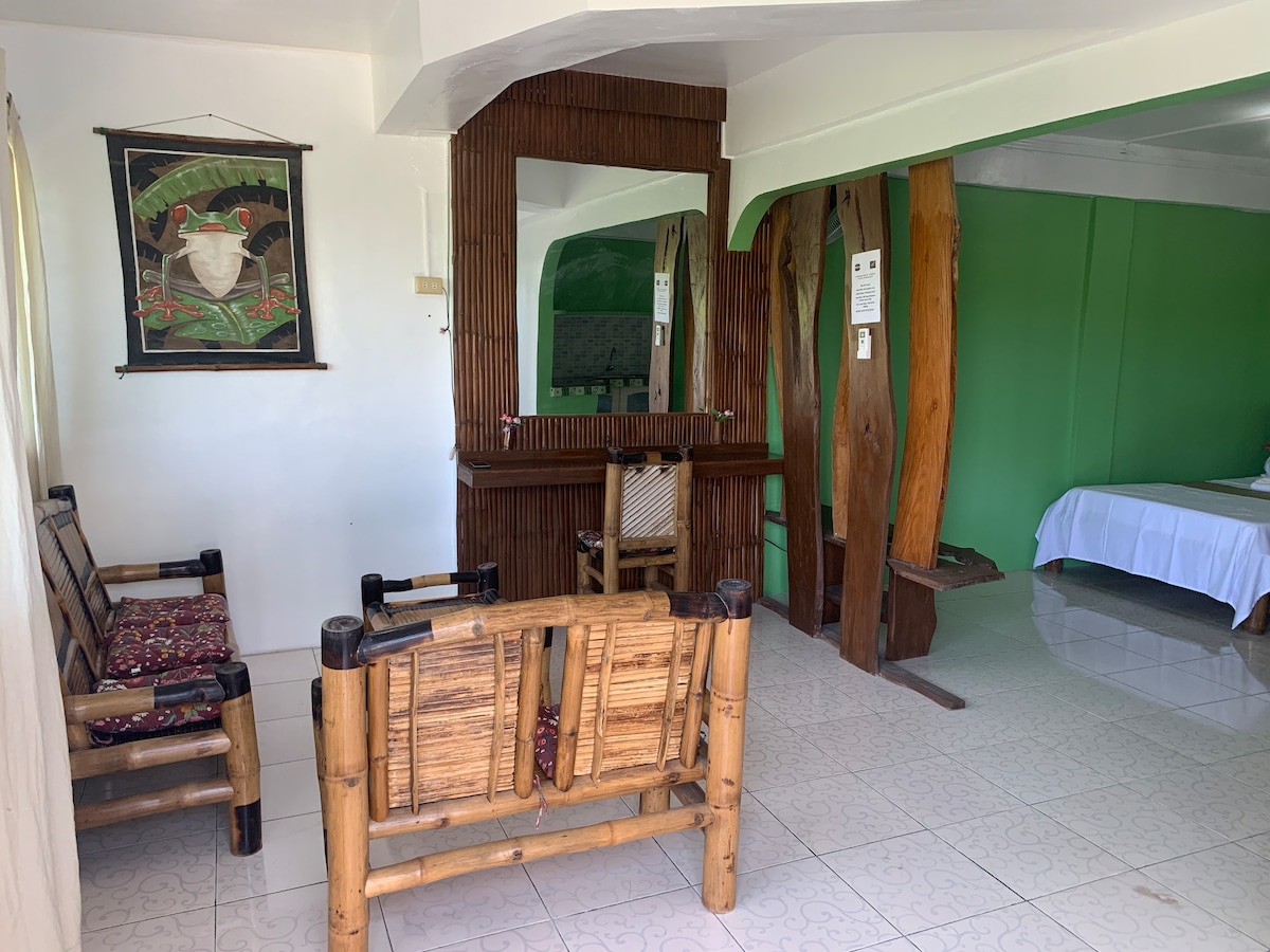 A seating area features bamboo chairs with patterned cushions, providing a casual space to relax. A large mirror reflects natural light, while a wooden table and decorative wall art enhance the room's comfort. The nearby sleeping area is separated by textured wooden elements.