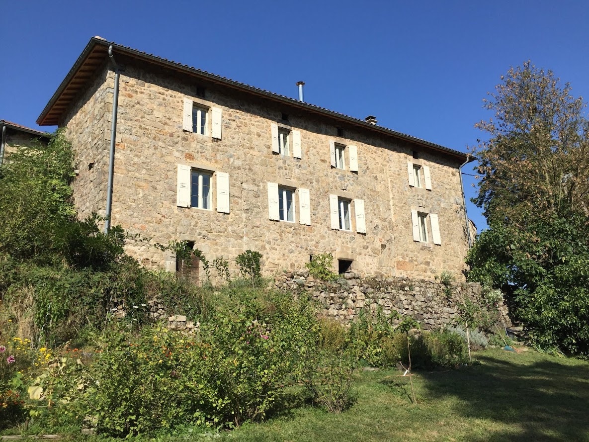 The sturdy stone house is set against a clear blue sky, featuring multiple windows framed by wooden shutters. Lush greenery surrounds the property, and a well-maintained garden is visible in the foreground, highlighting the natural beauty of the area.