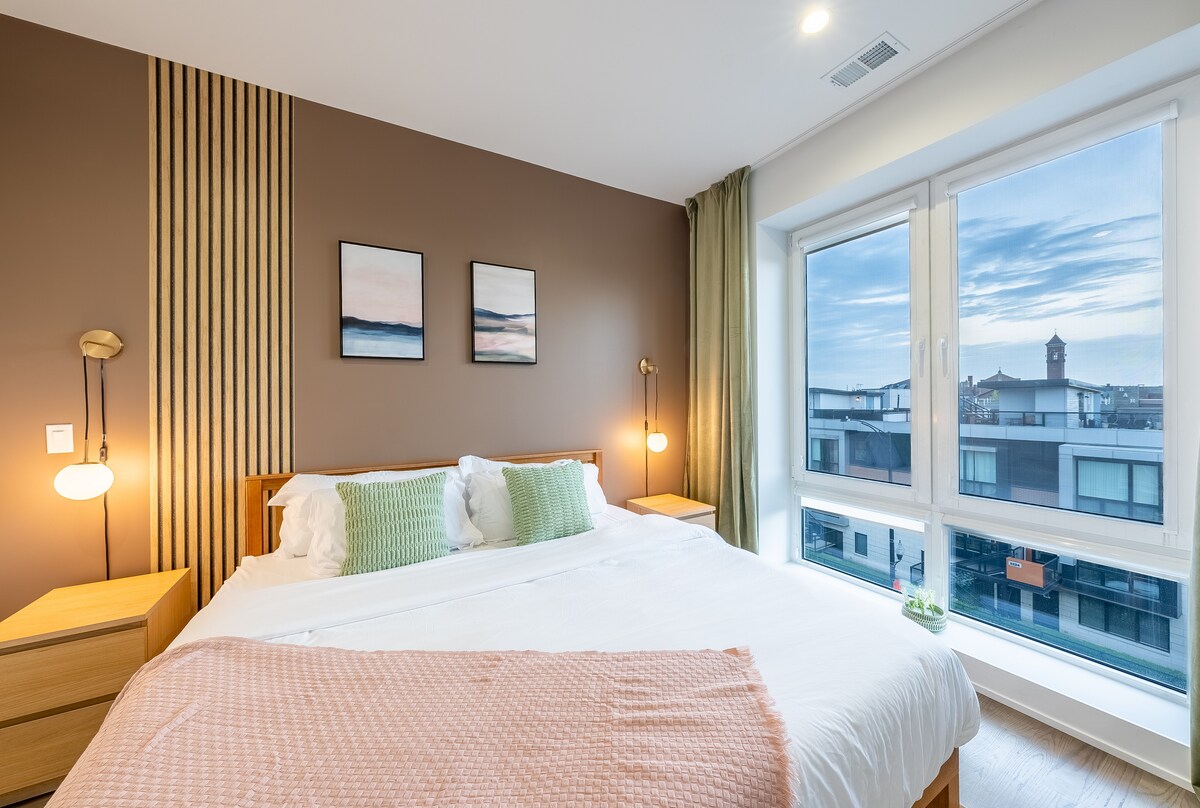 A well-appointed bedroom features a spacious bed with soft white linens, complemented by decorative pillows. Two nightstands with elegant lamps flank the bed, while large windows offer a view of the outdoors, inviting natural light into the room.