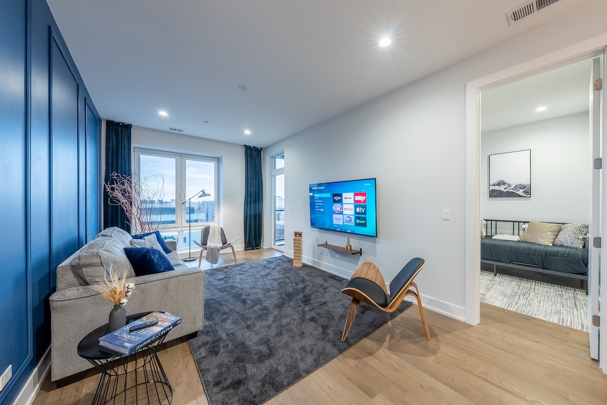 A modern living room features a plush gray sofa and an elegant armchair, positioned on a soft area rug. A large TV is mounted on the wall, with a view of large windows allowing natural light. Light-colored walls complement the blue accent wall.