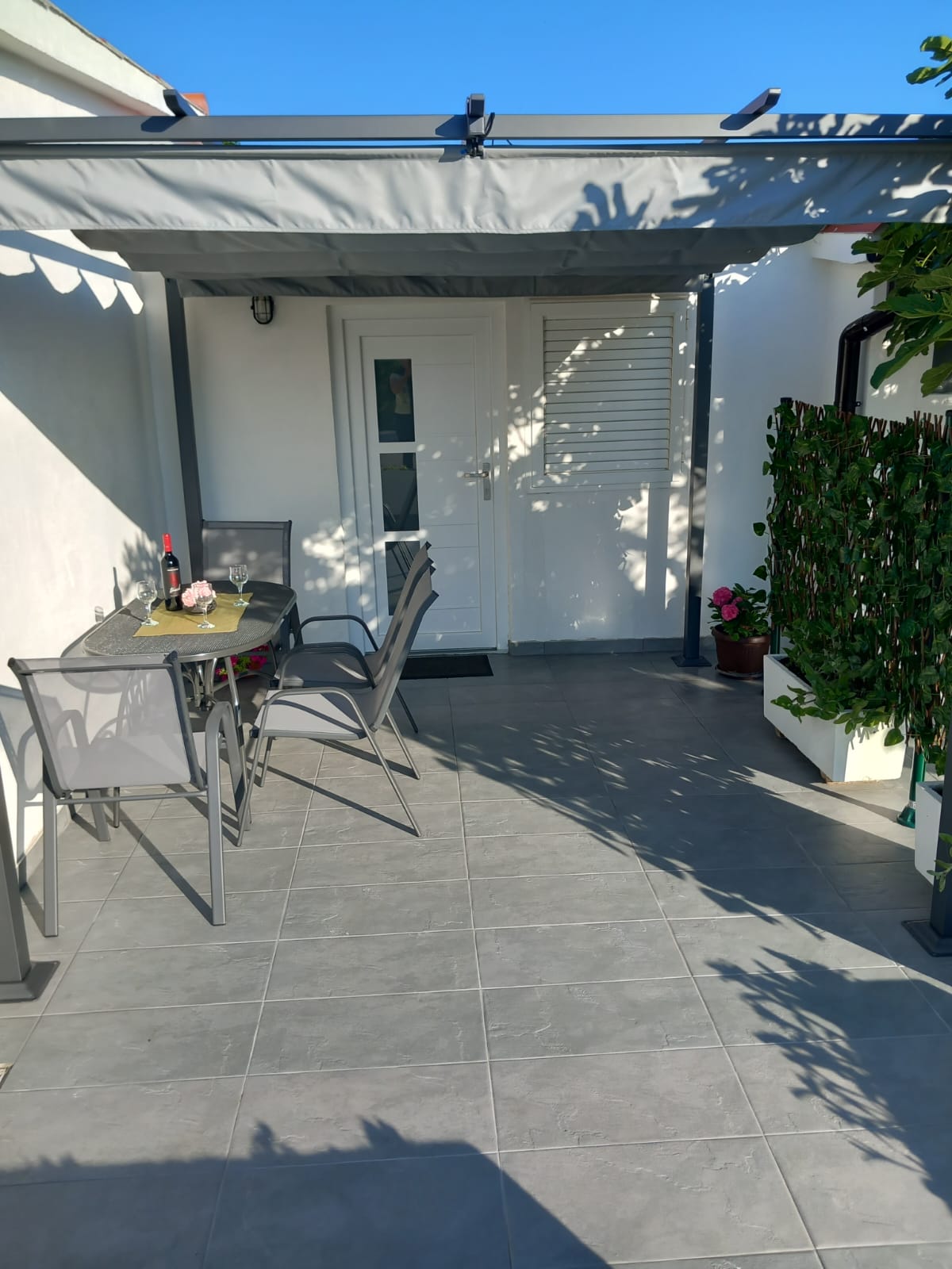 A covered patio is featured, furnished with a table and chairs suitable for outdoor dining. The space is paved with gray tiles, and a small plant is positioned at the side, contributing to a serene setting shaded by the overhead awning.
