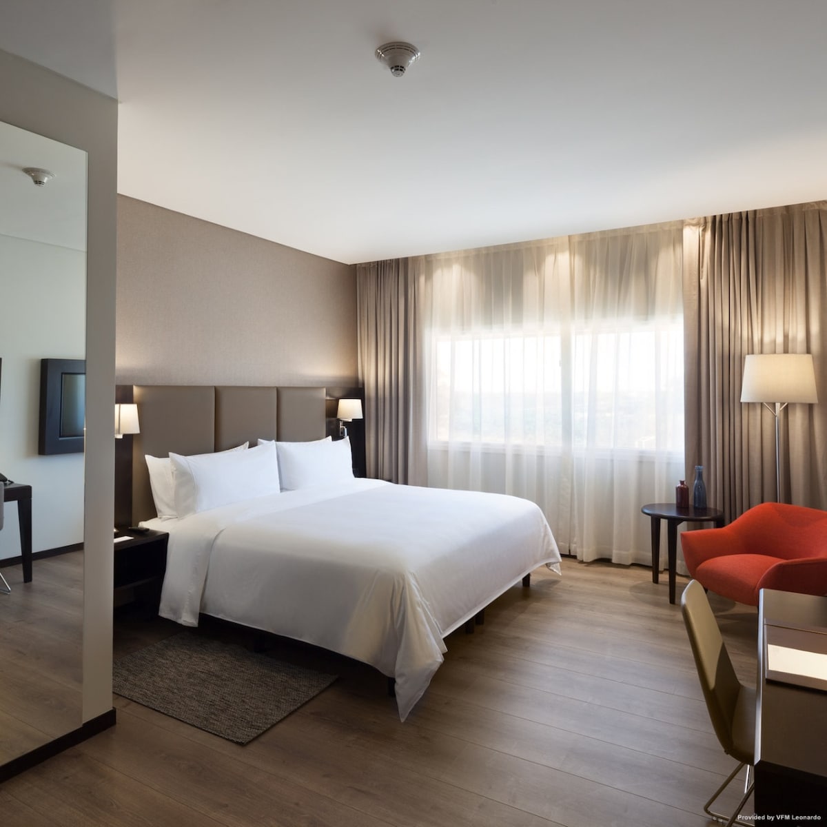 A modern hotel suite is depicted, featuring a spacious bed with crisp white linens and a decorative headboard. Natural light filters through sheer curtains over a large window. A small work desk and a lounge chair are visible, contributing to a functional and comfortable atmosphere.