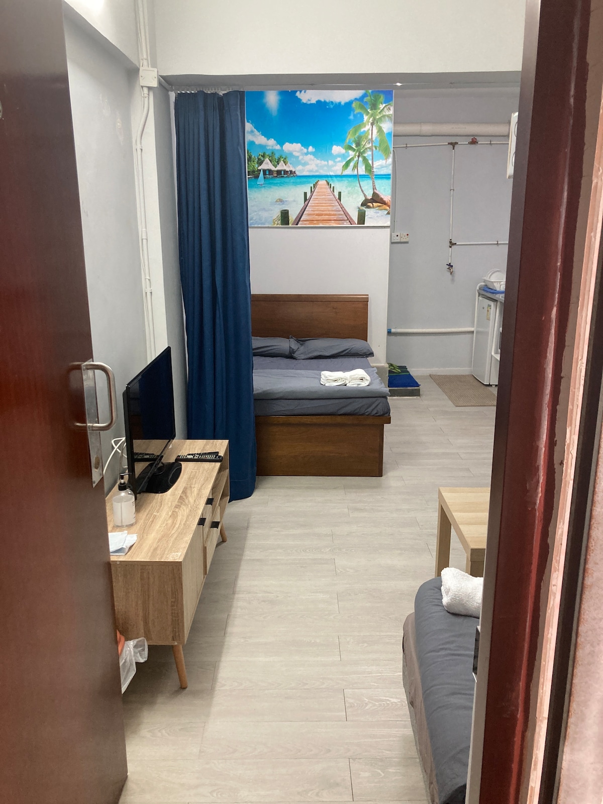 A cozy room features a wooden bed, a small sofa, and a light wooden table. A vibrant mural of a tropical scene decorates the wall, while a dark curtain provides privacy. A laundry area is visible in the background, contributing to the space's functionality.