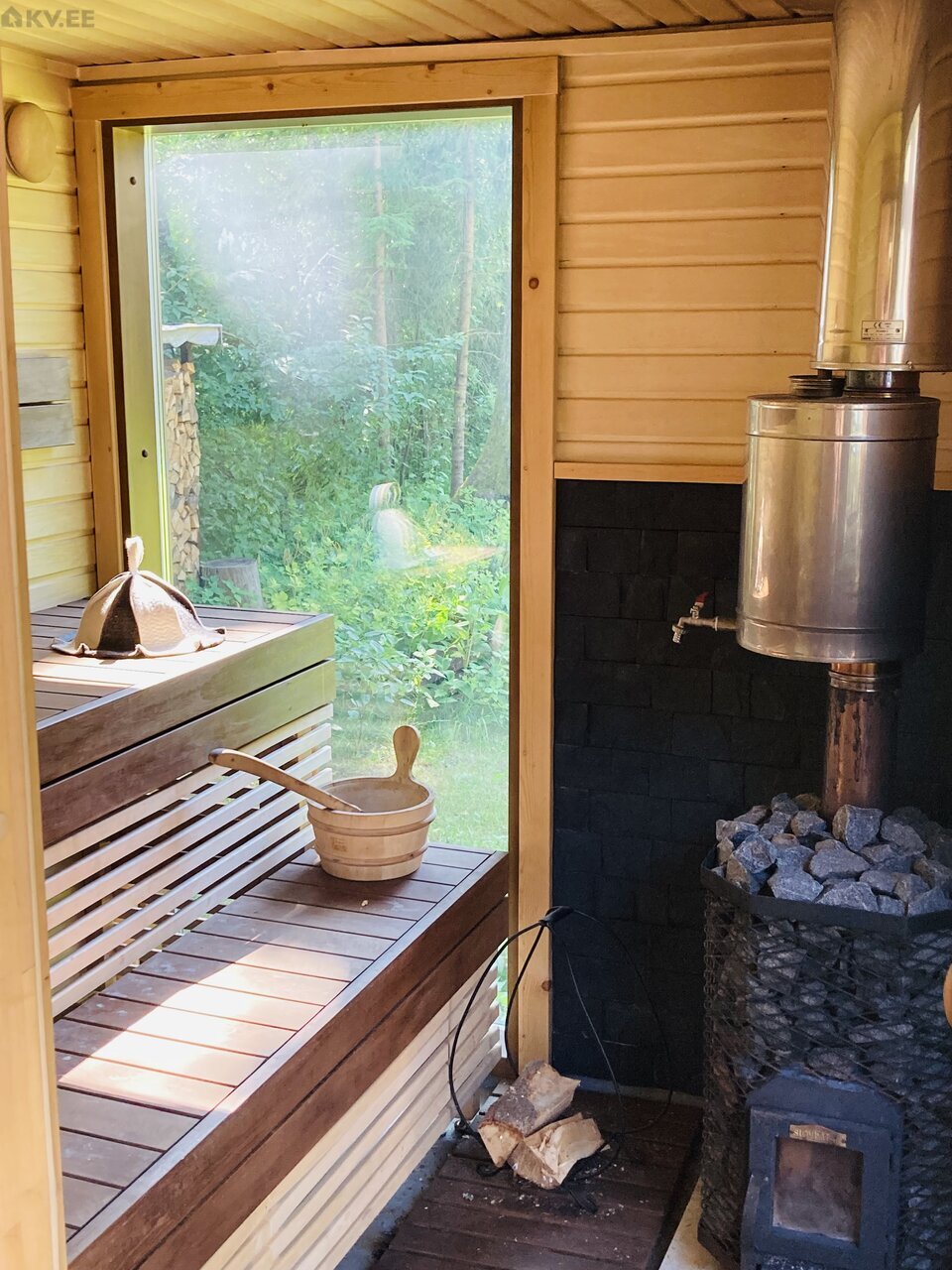 The sauna features wooden benches crafted from slats against a backdrop of natural wood walls. A large window offers views of the surrounding greenery. A traditional sauna stove is positioned beside stacked stones, while a wooden bucket and ladle are neatly placed on the bench.
