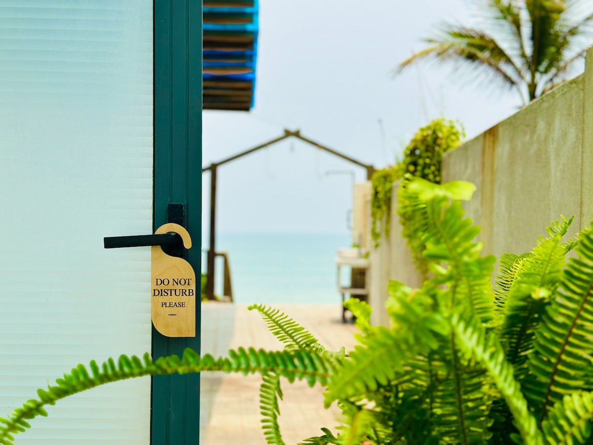 A door with a wooden 'Do Not Disturb' sign is framed by lush green ferns, guiding the view towards a pathway leading to the beach. The calm sea and distant structure are visible through an opening, enhancing the sense of tranquility.