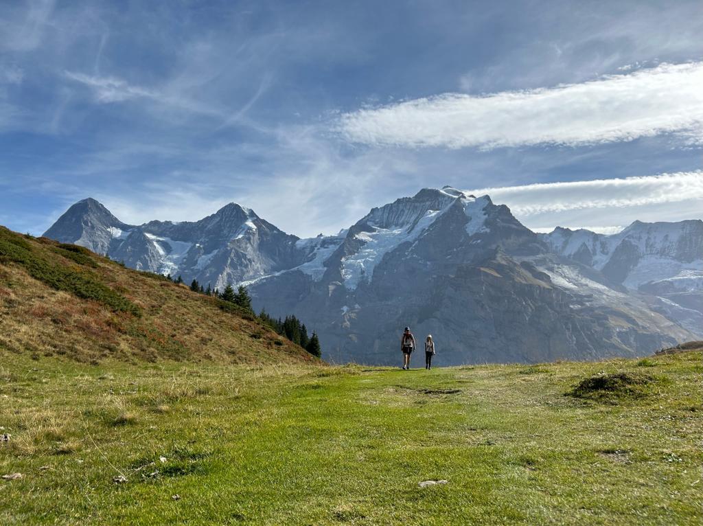 A scenic outdoor view showcases two hikers walking along a grassy path, set against a backdrop of majestic mountains under a clear blue sky. The landscape reflects the natural beauty of the surrounding area, emphasizing the tranquility and allure of the hiking experience.