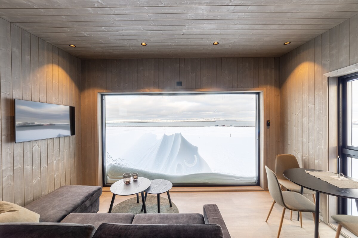 A modern living area features a large window offering scenic views of a snow-covered landscape. A cozy seating arrangement with a dark sofa and two small side tables sits before the window. Natural light fills the space, highlighting the warm wooden interior.