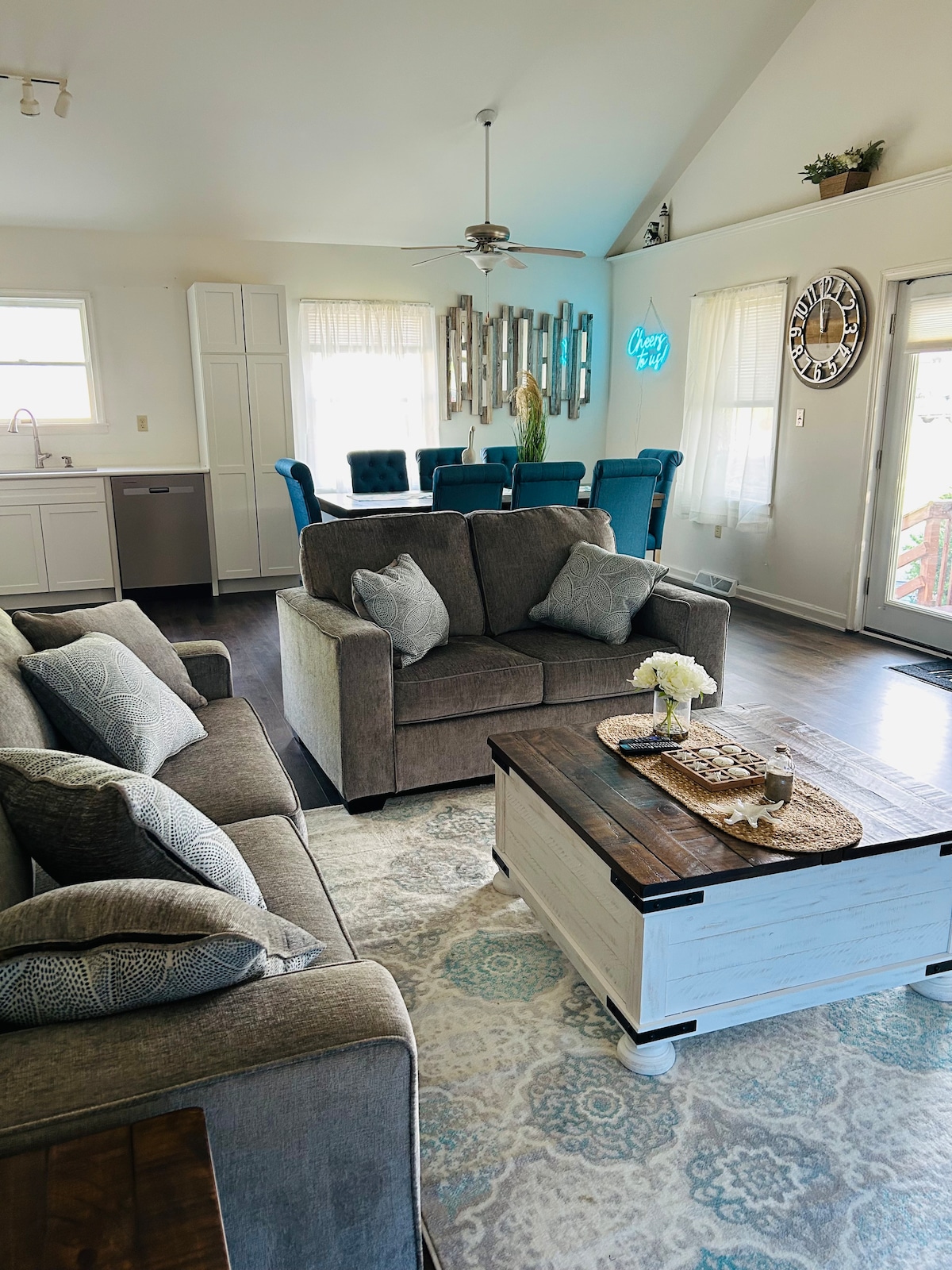 The living area showcases a blend of comfortable seating with a gray sofa and armchairs, complemented by decorative pillows. A light-colored coffee table sits at the center, adorned with a floral arrangement. The dining area is visible, featuring a long table surrounded by blue chairs.