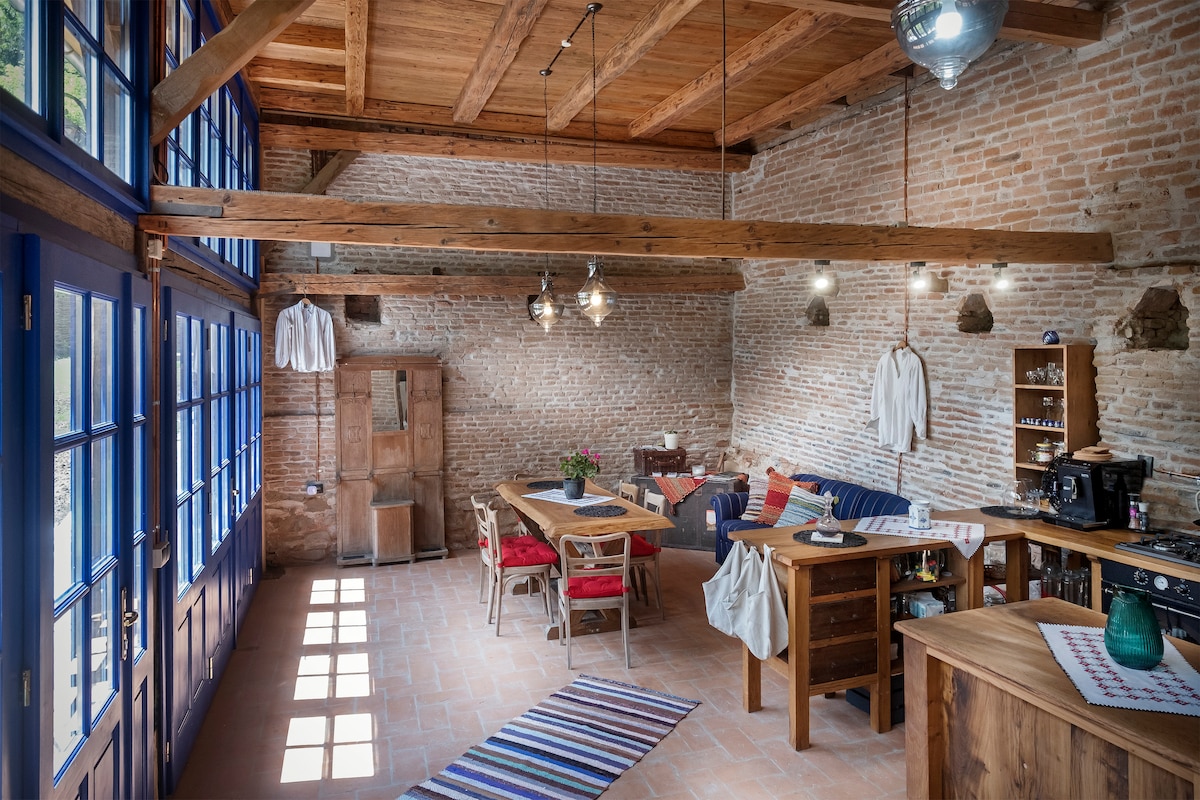 The open space kitchen and dining area features rustic brick walls and wooden beams. Sunlight filters through large blue-framed windows. A solid wood dining table is surrounded by colorful chairs, and the kitchen area includes wooden countertops and modern appliances, creating a warm and inviting atmosphere.