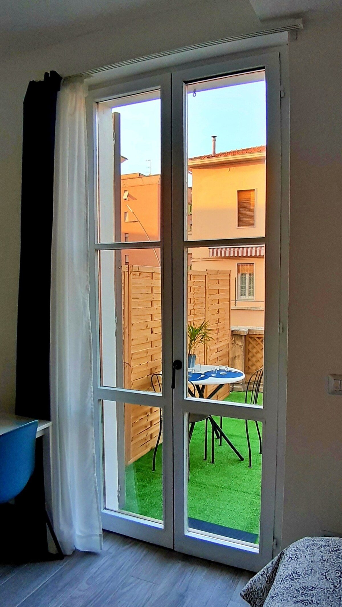 A large window offers a view of a terrace with a small table and chairs. Light streams in, enhancing the space. Greenery and wooden fencing create a cozy outdoor setting, while the interior is complemented by a workspace and light-colored curtains.