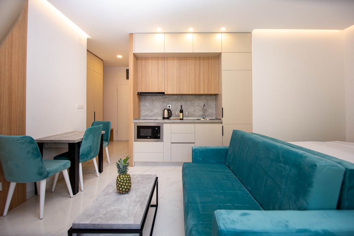 A modern interior features a cozy seating area with a teal sofa and a small coffee table. A dining space with four upholstered chairs is visible. The kitchenette is equipped with essential appliances and cabinetry, highlighted by a neutral color palette and warm wooden accents.