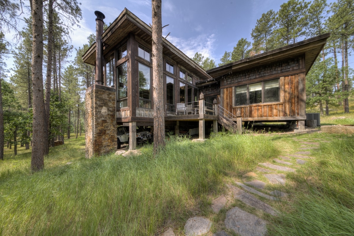 A unique lodge is nestled among towering trees, featuring a blend of wood and glass in its design. A stone pathway leads to the entrance, while expansive windows provide a view of the natural surroundings. The setting showcases a harmonious connection with nature.