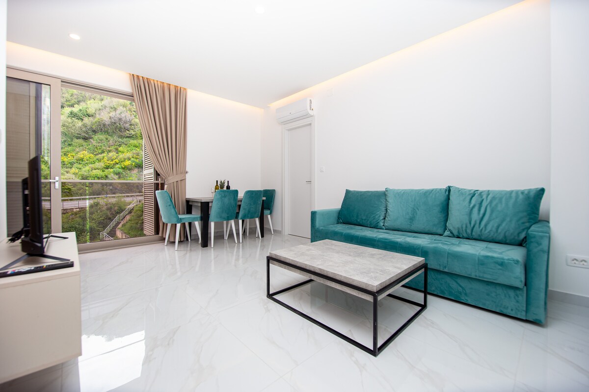 A modern living area features a plush turquoise sofa and a sleek coffee table. Large glass doors provide natural light and a scenic view of lush greenery. A dining table with teal chairs complements the bright and airy atmosphere.