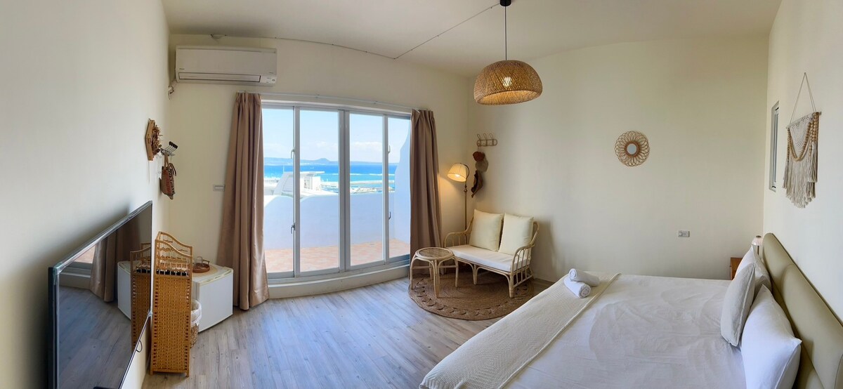A bright and airy bedroom features large glass doors that provide a view of the ocean. A spacious bed is complemented by neutral-colored furnishings. A comfortable seating area is positioned beside a decorative light fixture, while natural textures contribute to a calm and relaxed environment.
