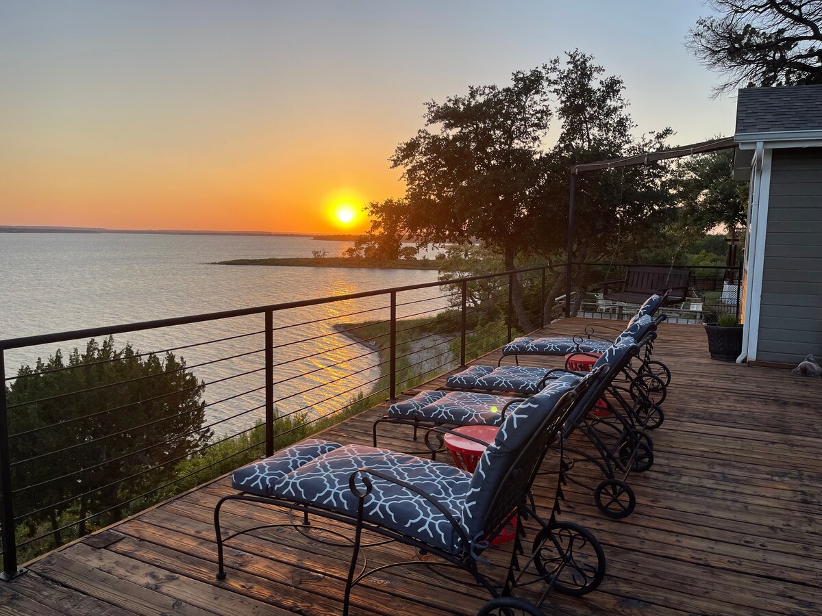 A spacious deck is equipped with multiple lounge chairs facing the lake, offering panoramic sunset views. The calm water reflects the orange and pink hues of the sunset, while trees frame the scene, creating a serene atmosphere.
