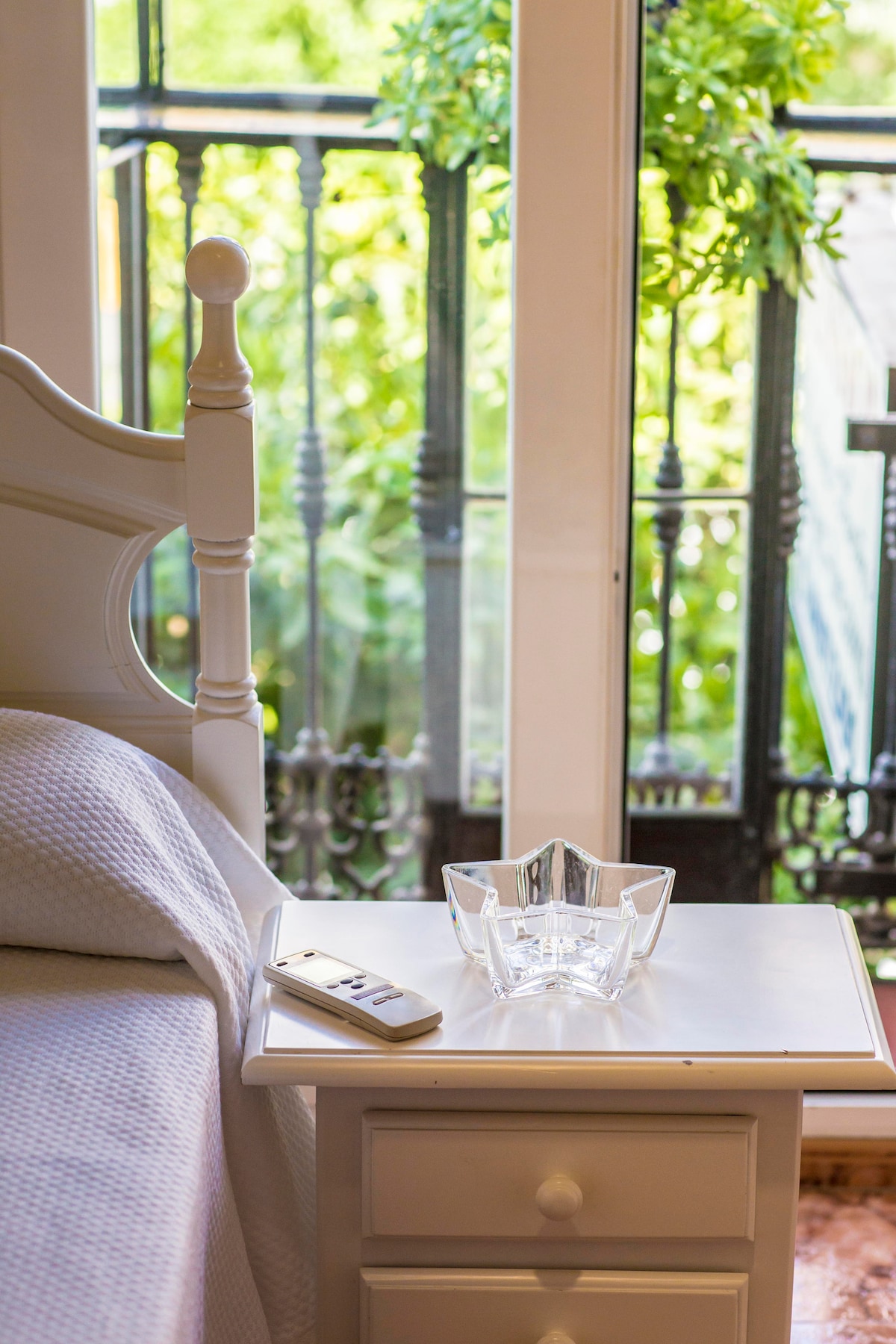 A bedside table features a crystal dish and a remote, positioned beside a spacious bed. A balcony with wrought iron railings is visible through the glass doors, framed by greenery that softly diffuses the light entering the room.