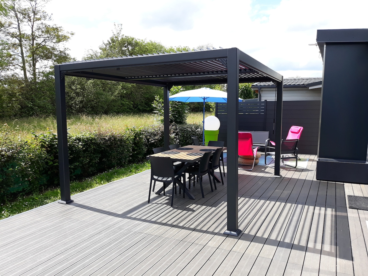 A covered outdoor seating area is featured, with a large table surrounded by six chairs. A colorful umbrella provides shade, while additional lounge chairs in vibrant hues are positioned nearby. Lush greenery borders the space, creating a secluded environment.