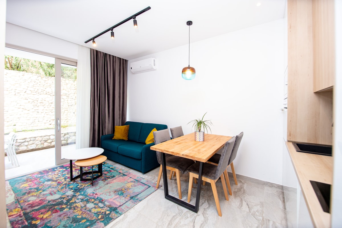 A living area features a blue sofa along with a wooden dining table surrounded by gray chairs. Natural light is provided through patio doors, and a colorful rug adds visual interest to the neutral-toned flooring.