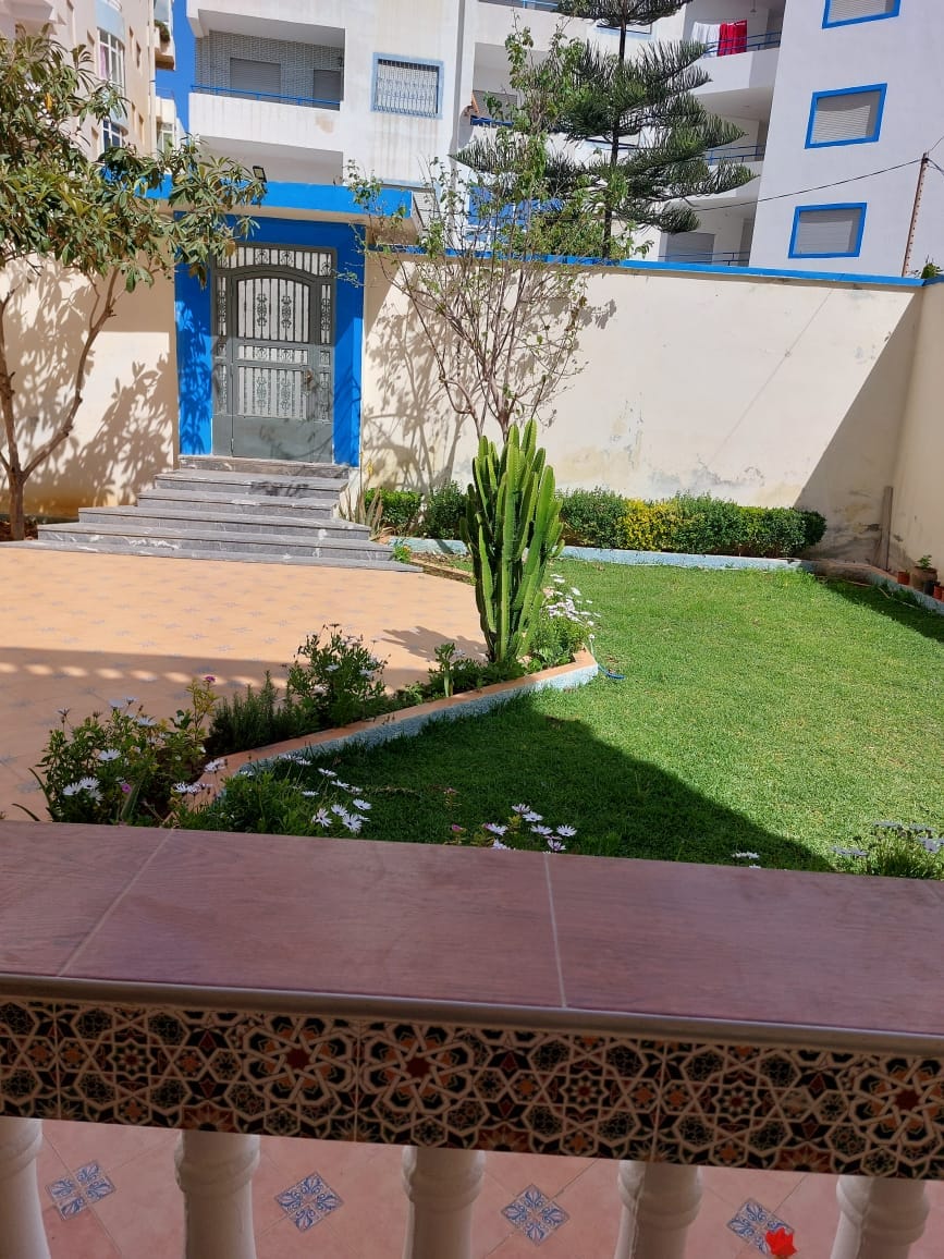 A well-kept garden area features lush green grass bordered by various plants and flowers. A stone pathway leads to a gated entrance. The image shows a view from a balcony, highlighting the serene outdoor space, with a gentle slope and neat landscaping.