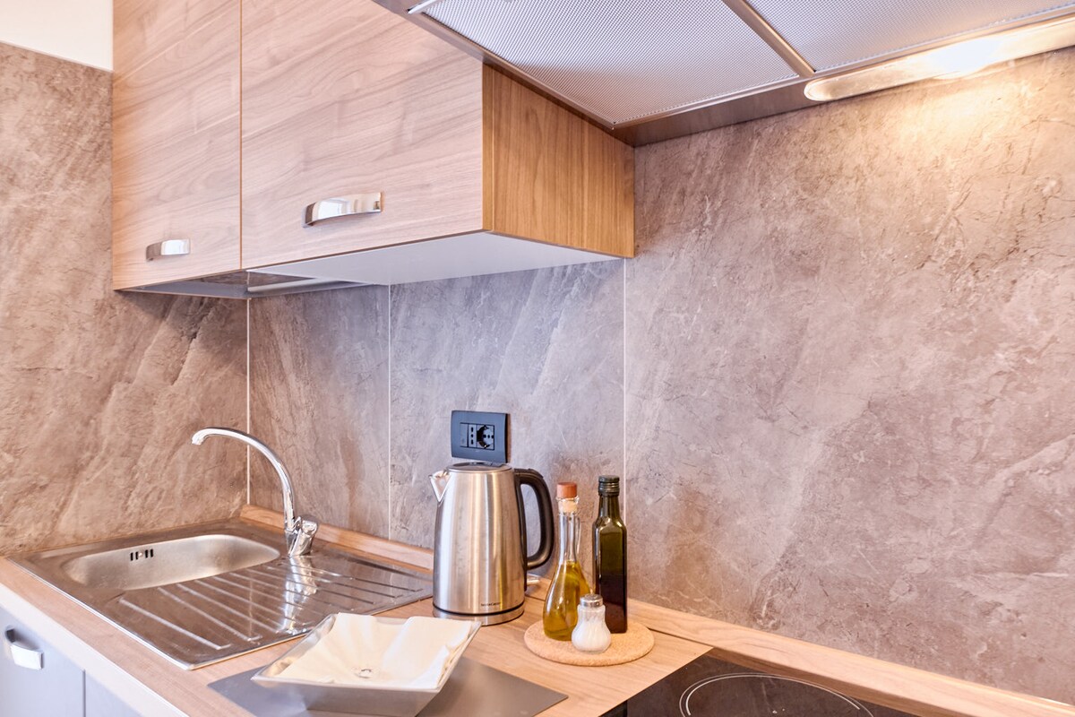 A modern kitchen area is showcased, featuring wooden cabinets and a sleek countertop. A kettle and oil dispenser are positioned next to a small bowl for food preparation. Additionally, an induction cooktop is visible, providing essential cooking amenities.
