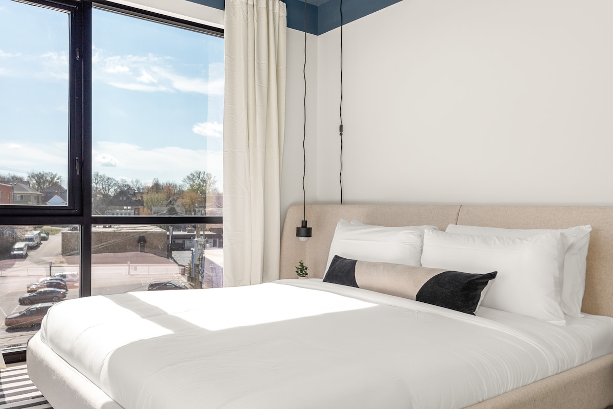 A spacious bedroom features a comfortable bed with crisp white linens and decorative pillows. Large windows allow natural light to brighten the room, offering views of the surrounding area. Minimalist decor enhances the clean and modern aesthetic.