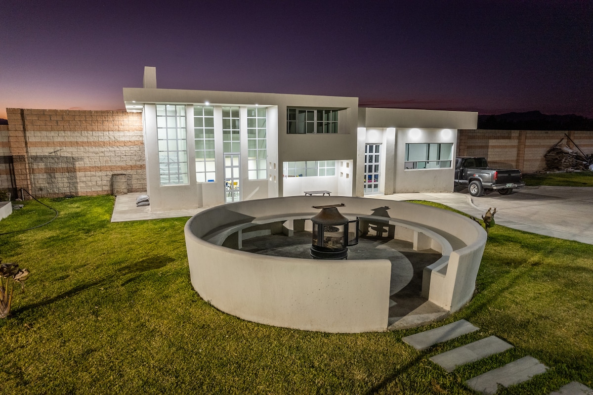 A modern building features large glass windows and a sleek, circular seating area surrounding a fire pit. The surrounding grassy area is well-maintained, leading to stone pathways and a spacious parking area, creating an inviting outdoor gathering space.