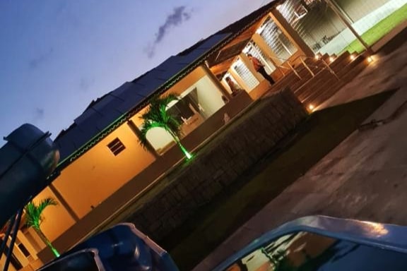 An exterior view of the house is presented at dusk, with strategic lighting highlighting the architecture. A smooth pathway leads towards the entrance, adorned with palm trees. The gentle glow from the house enhances the serene atmosphere, suggesting a comfortable space for relaxation.