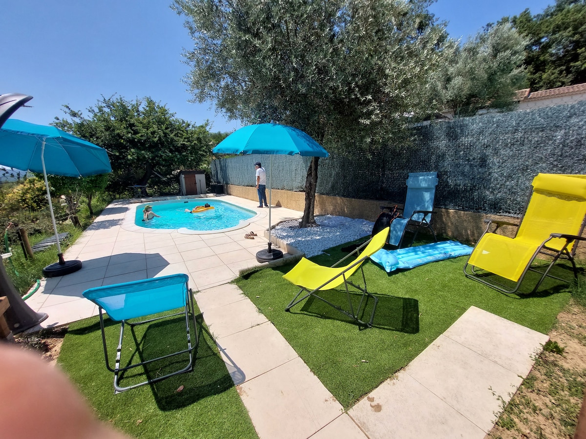 The outdoor space features a securely fenced swimming pool surrounded by lounge chairs and umbrellas for shade. A grassy area complements the paved patio, with lush greenery visible in the background, offering a relaxing atmosphere for guests.