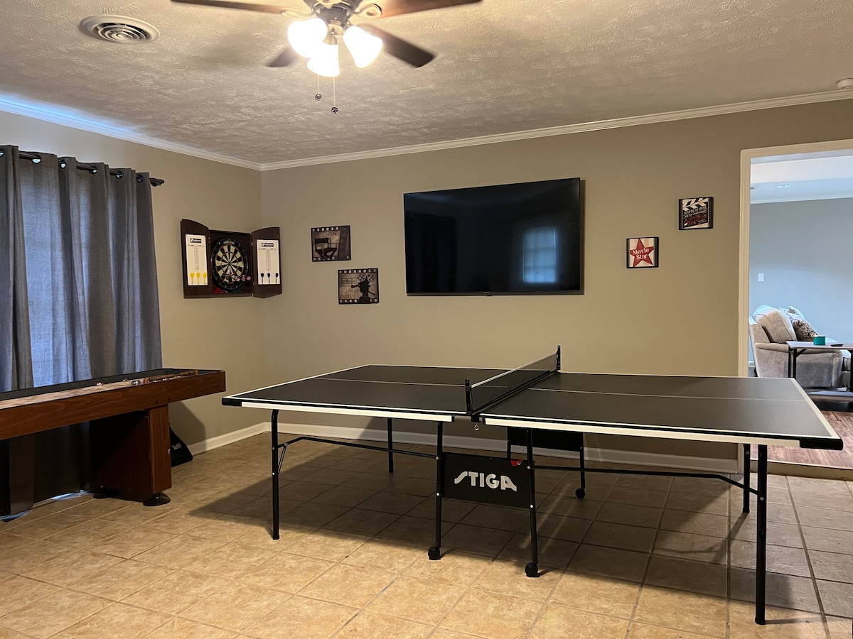 An entertainment area features a black ping pong table positioned centrally. A large flatscreen TV is mounted on the wall, above a wooden dartboard and game scoreboards. Light-colored walls are complemented by dark curtains, and a light ceiling fan is present.