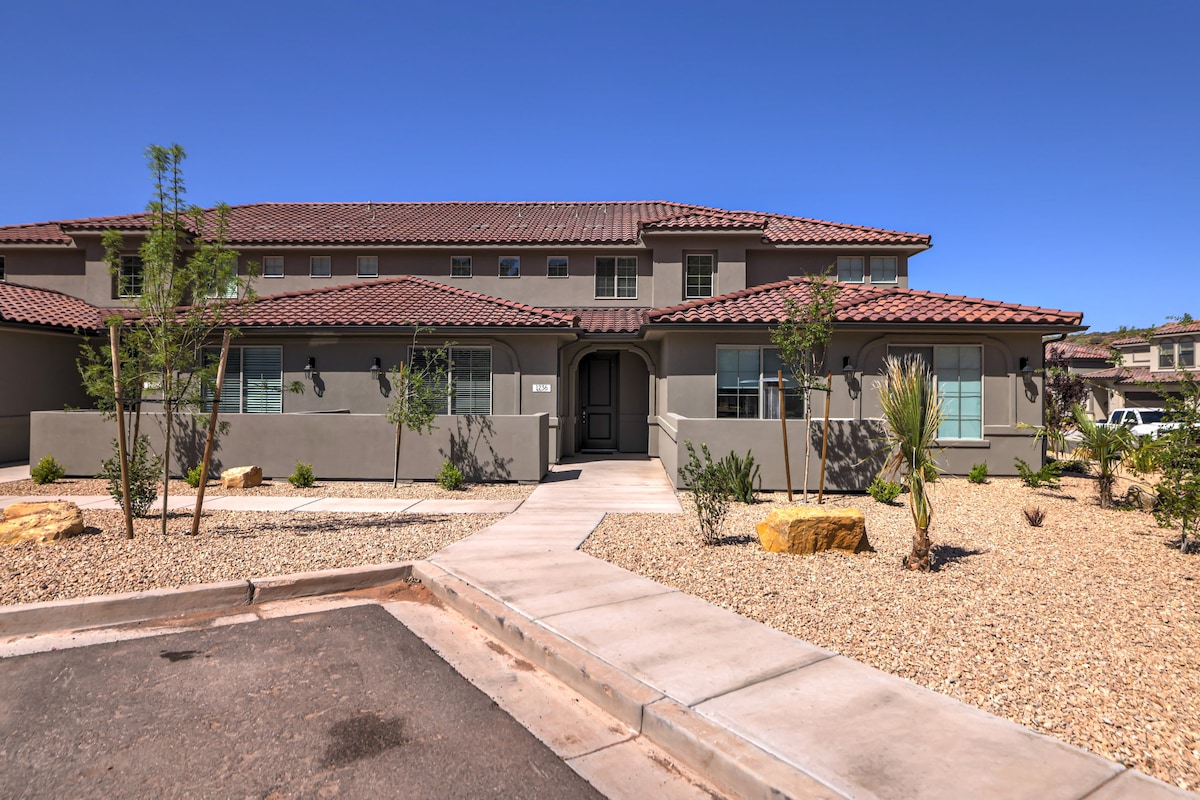 The exterior of a modern townhouse showcases a landscaped area with gravel and desert plants. A paved walkway leads to the entrance, framed by large windows on either side. The two-story structure is adorned with a tile roof and neutral-toned walls, complemented by clear blue skies.