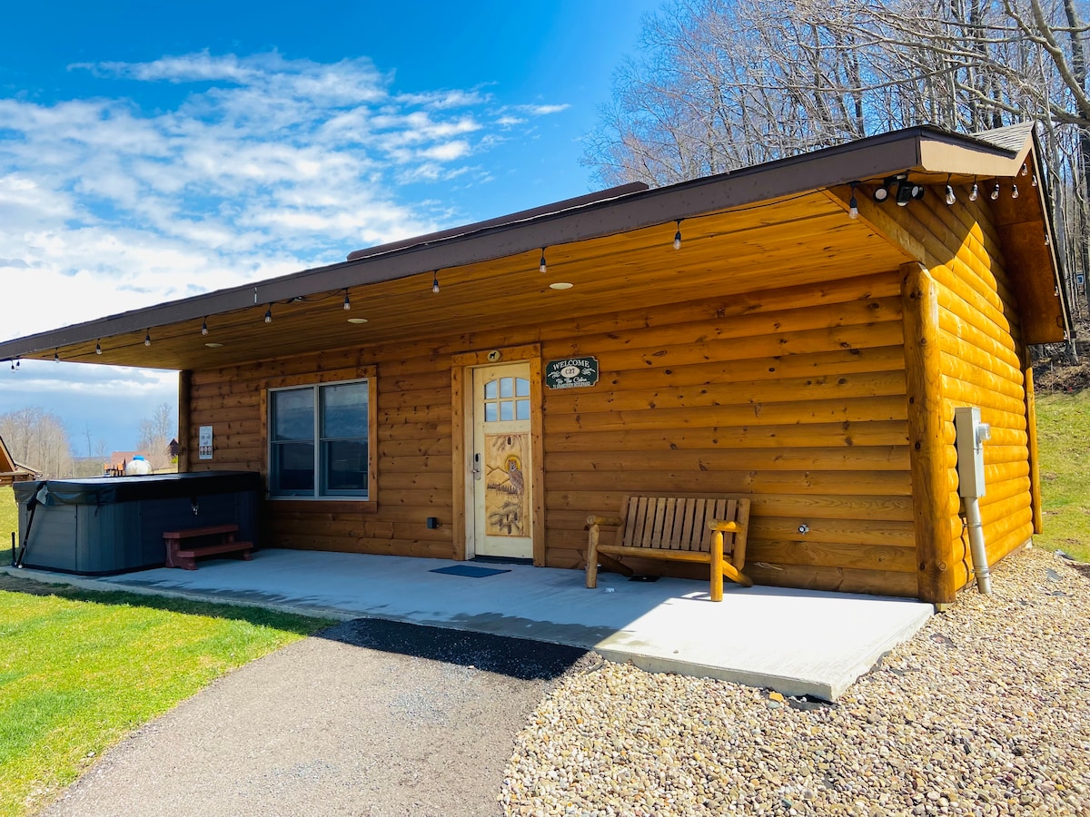 A log cabin exterior features a warm wooden facade with a welcoming entrance. A bench is situated next to the door, and a hot tub is positioned on the side. A spacious outdoor area with green grass complements the rustic charm of the structure.