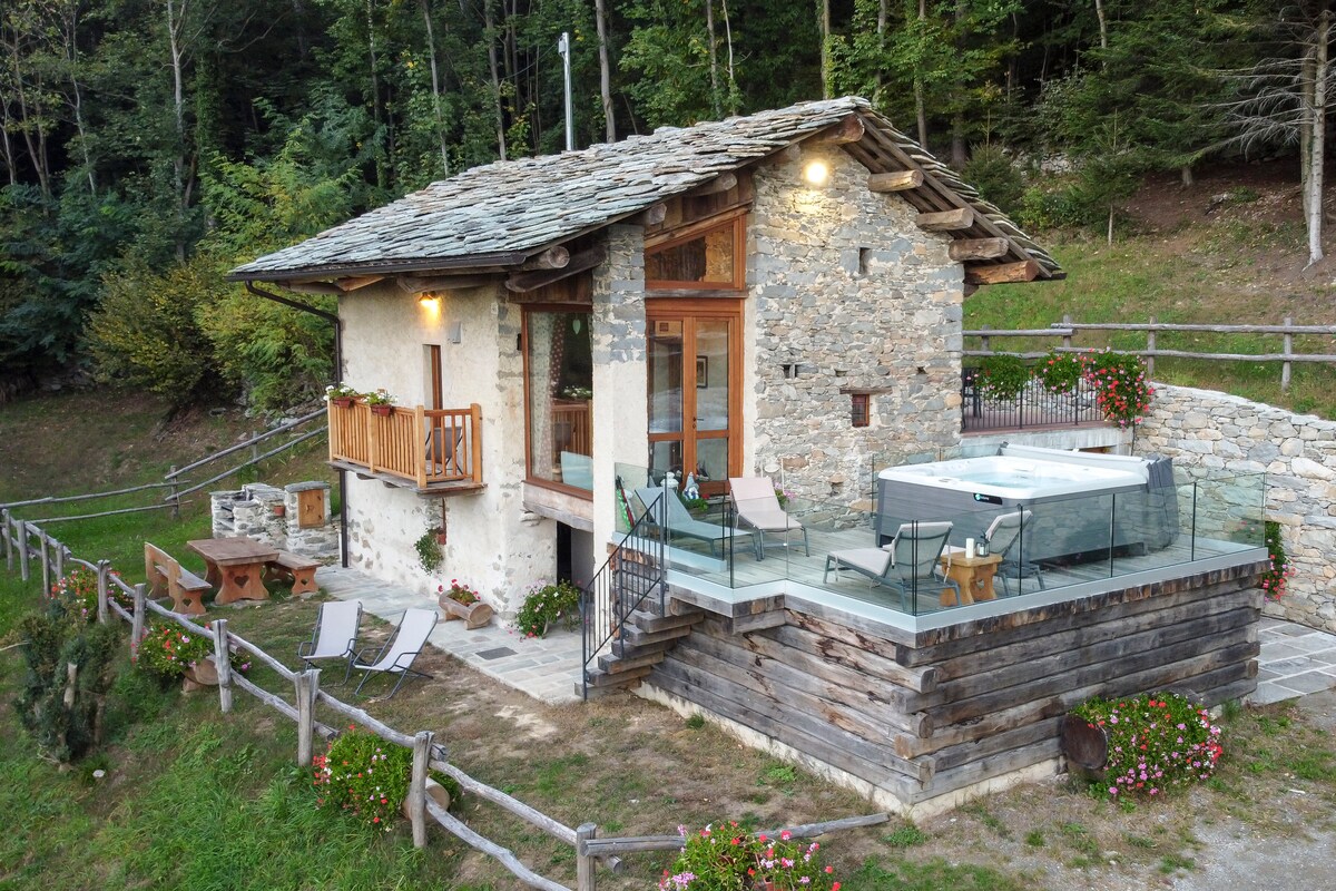 A rustic chalet constructed from natural stone and wood is surrounded by greenery. A spacious terrace features a hot tub and outdoor seating. Large windows and a balcony enhance the serene outdoor views. The inviting garden is adorned with flowers and bordered by a wooden fence.