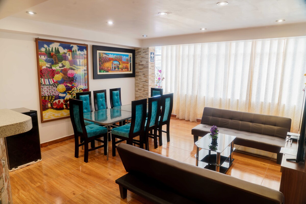 A spacious living and dining area features a glass dining table surrounded by six upholstered chairs in a vibrant blue hue. A comfortable brown sofa complements a glass coffee table, with artwork on the walls adding color. Bright natural light filters through sheer curtains.