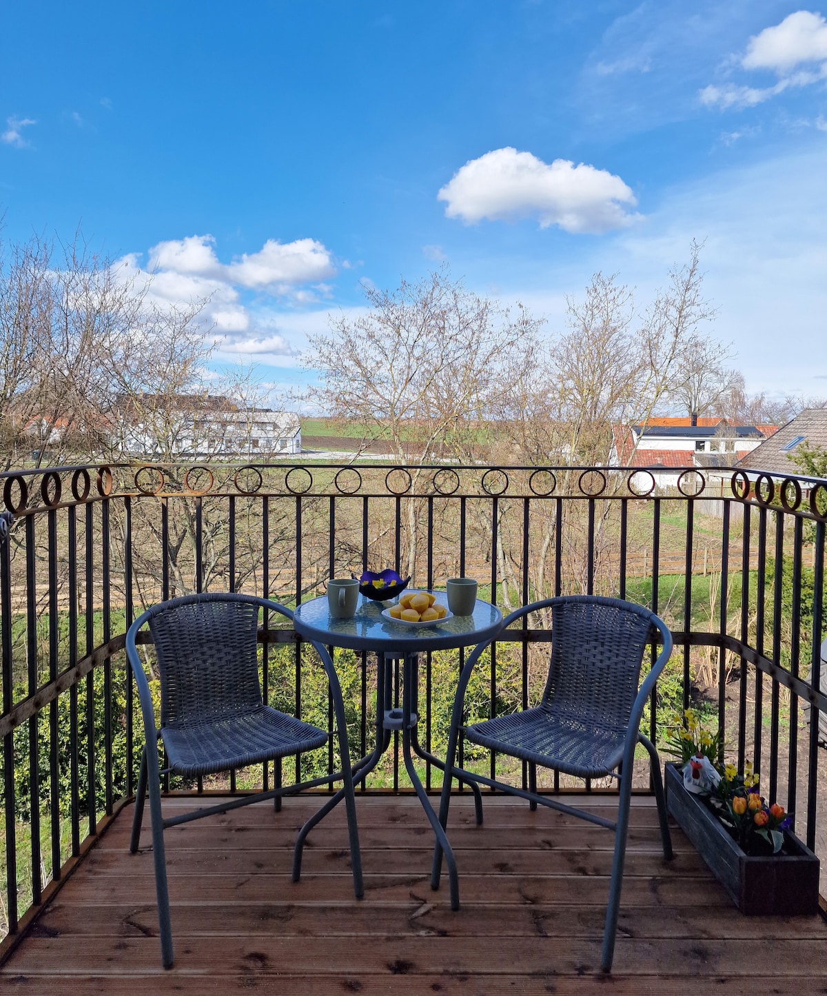 A small outdoor balcony is shown with a round table and two chairs, overlooking a green landscape. A decorative flower box sits nearby, and the sky is bright with fluffy clouds. Fresh fruits are displayed on the table, creating a casual dining space.