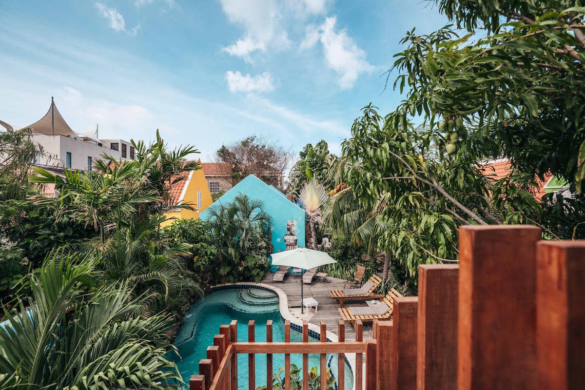 A serene tropical garden surrounds a refreshing pool, which is shaded by an umbrella. Vibrant blue and yellow buildings are partially visible through lush greenery, contributing to a tranquil atmosphere in the heart of the vibrant Pietermaai area.