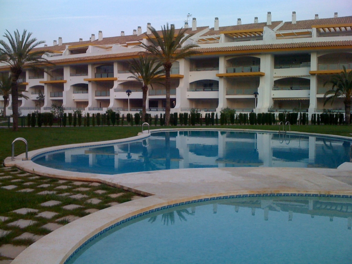 Two inviting swimming pools, surrounded by well-maintained gardens, reflect the soft light of the day. The building in the background features multiple terraces and is framed by palm trees, creating a serene setting for relaxation.