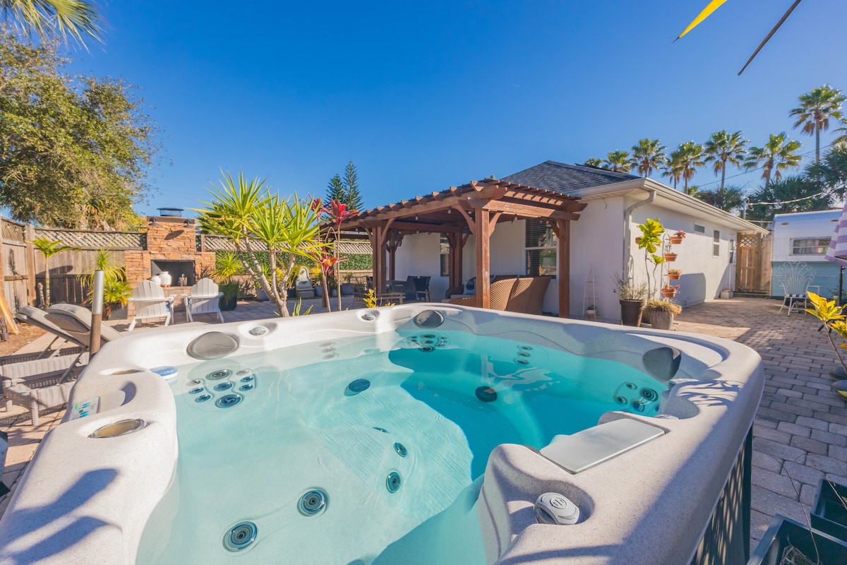 A large hot tub is centrally located in a spacious backyard, surrounded by palm trees and vibrant plants. An outdoor seating area is visible in the background, along with a well-maintained facade of the house. Clear blue skies enhance the inviting atmosphere.