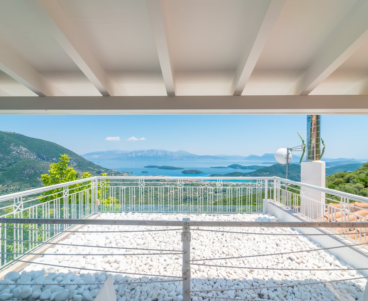 A spacious terrace is highlighted, featuring a white gravel surface and a railing. The panoramic views of the surrounding mountains and the blue waters of the sea create a serene backdrop, complemented by the clear sky.