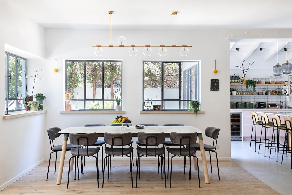 A modern dining area features a long table surrounded by ten chairs. Two large windows allow natural light to flood the space, with greenery visible outside. The kitchen area can be seen in the background, showcasing organized shelves and appliances.