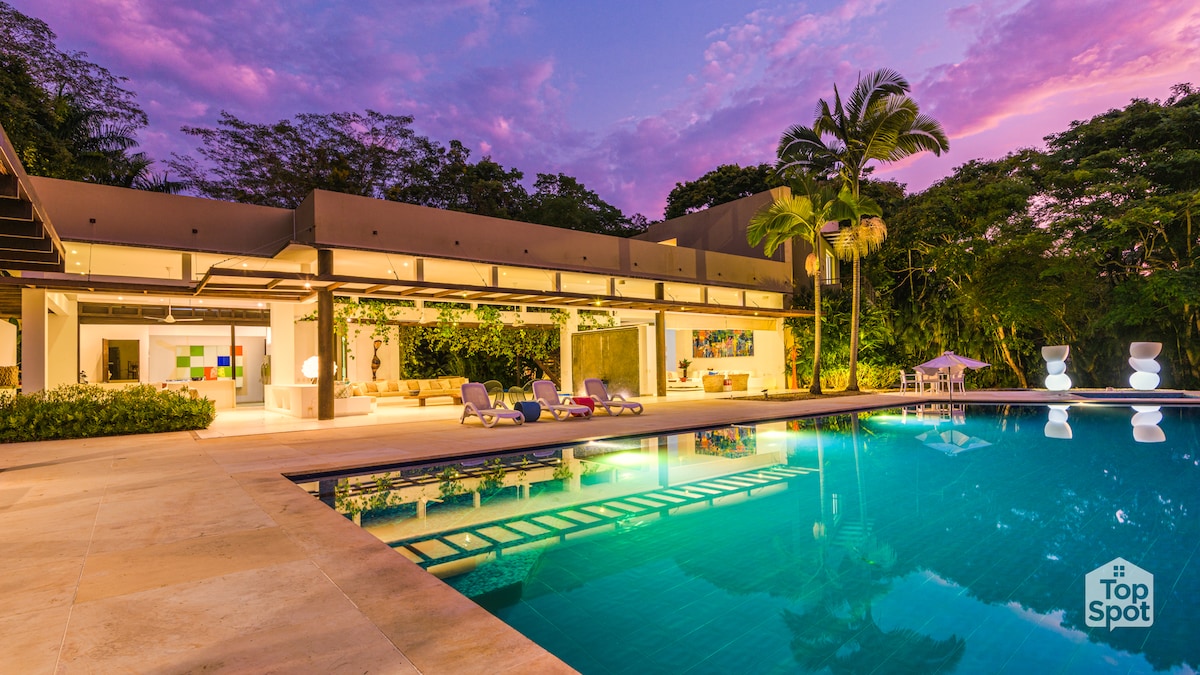 A modern two-story property is set against a vibrant sunset sky. The inviting pool reflects colorful clouds, while loungers are positioned nearby. Lush greenery surrounds the outdoor space, enhancing the tranquil atmosphere. Large glass doors provide a view into an open living area that blends indoor and outdoor environments.