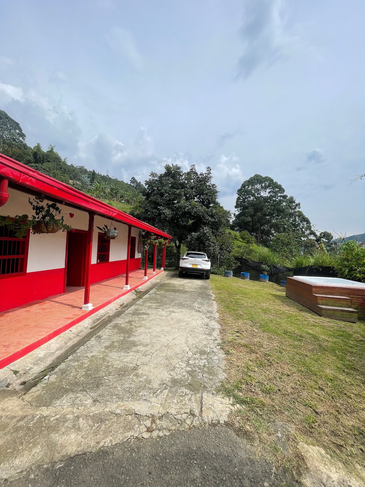 A red-walled building extends along a stone path, leading to a grassy area. A white car is parked nearby, with trees and blue barrels visible in the background. The sky is partly cloudy, enhancing the serene outdoor setting.